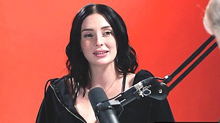 Charlotte Sins talks into mic in black hoodie against red studio wall